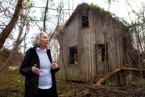 Older woman standing in front of a broken down wood house structure.